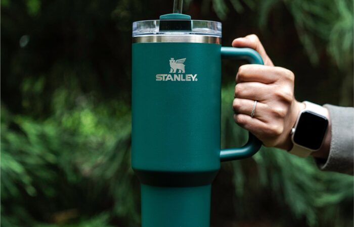This image features a person's hand holding a teal-colored Stanley Quencher H2.0​ Flowstate Tumbler. The mug has a prominent white Stanley logo on the front. The mug is equipped with a handle on the side and a straw protruding from the lid. The person is wearing a gray sleeve and a smartwatch on their wrist. The background is a blurred natural setting, suggesting the photo was taken outdoors, likely in a wooded area.