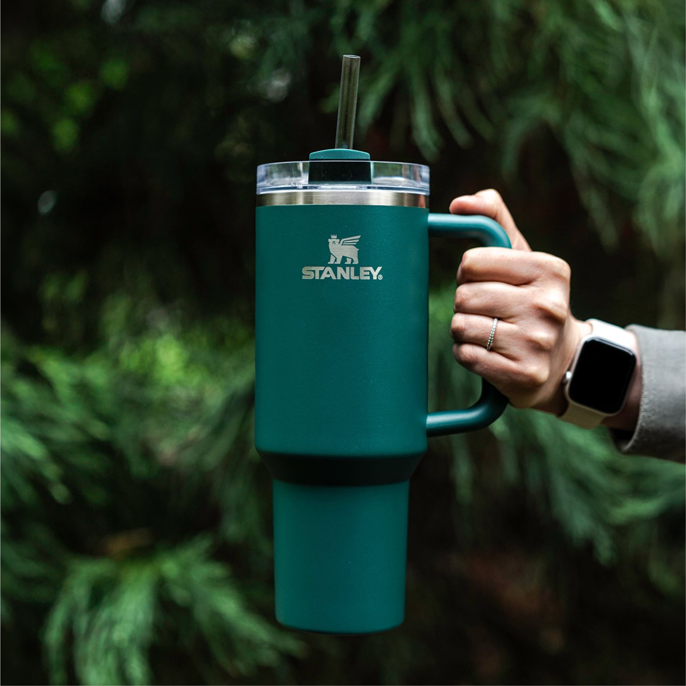 This image features a person's hand holding a teal-colored Stanley Quencher H2.0 Flowstate Tumbler. The mug has a prominent white Stanley logo on the front. The mug is equipped with a handle on the side and a straw protruding from the lid. The person is wearing a gray sleeve and a smartwatch on their wrist. The background is a blurred natural setting, suggesting the photo was taken outdoors, likely in a wooded area.